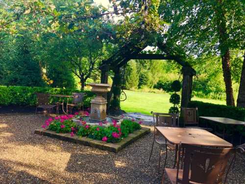 A serene garden scene with a stone fountain, colorful flowers, and metal tables under a wooden archway surrounded by greenery.