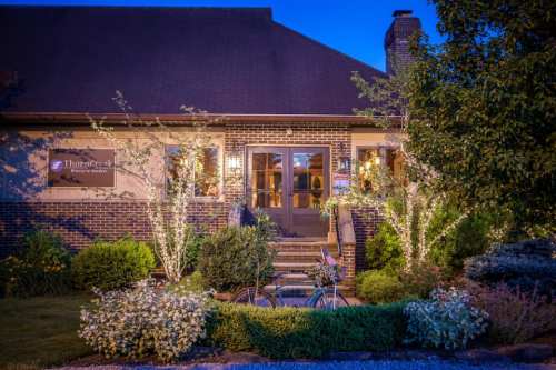A charming brick building with warm lights, surrounded by greenery and flowers, at dusk. A bicycle is parked in front.