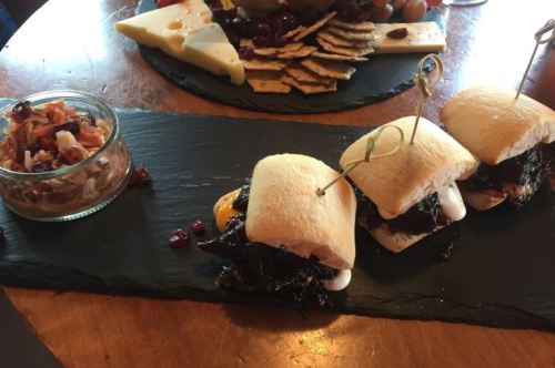 Three small sandwiches on a slate platter, accompanied by a bowl of chopped relish and a cheese platter in the background.