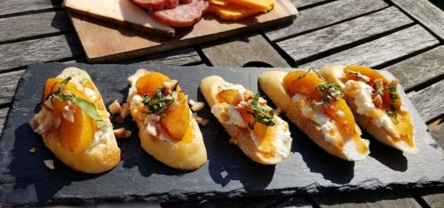 A slate platter with five crostini topped with peaches, cheese, nuts, and herbs, alongside a wooden board of meats and cheese.