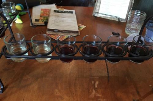 A flight of six small glasses filled with different beverages on a wooden table, with menus in the background.