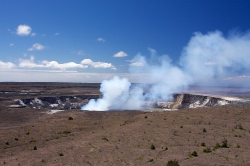 A volcanic crater with smoke rising against a blue sky and scattered clouds, surrounded by barren land.