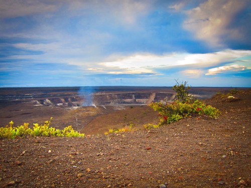 A panoramic view of a volcanic crater with smoke rising, surrounded by a barren landscape and a cloudy sky.