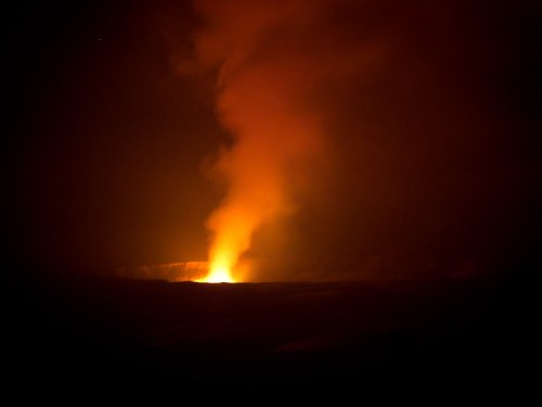 A glowing volcanic eruption emits a plume of orange smoke against a dark night sky.