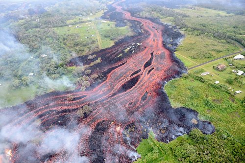 Aerial view of flowing lava creating a winding path through green landscape, with smoke rising in the background.