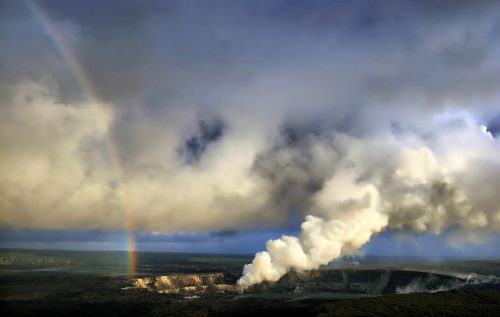 A volcanic landscape with steam rising, under a cloudy sky and a rainbow in the background.