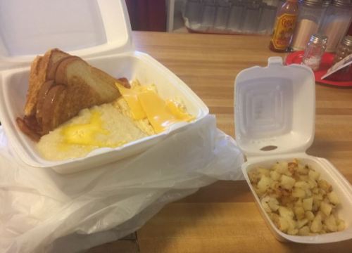 A takeout container with toast, cheese, eggs, grits, and a side of diced potatoes on a wooden table.