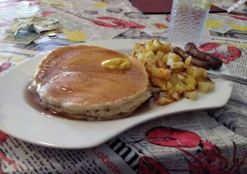 A plate of fluffy pancakes with butter and syrup, served with diced potatoes and sausage links on a patterned tablecloth.