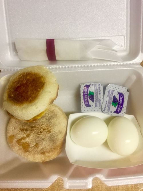 A takeout container with two English muffins, two boiled eggs, and grape jelly packets.