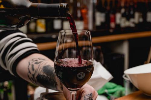 A hand pours red wine into a glass, with a blurred bar background and tattoos visible on the arm.