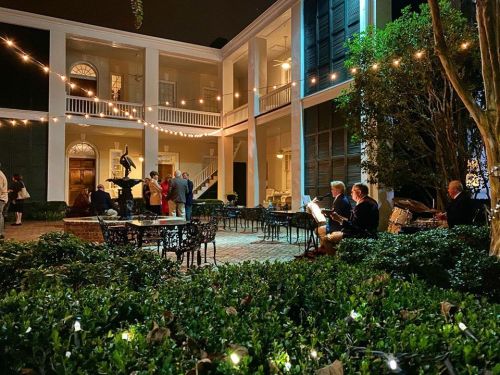 A nighttime courtyard scene with string lights, guests mingling, and musicians playing near lush greenery.