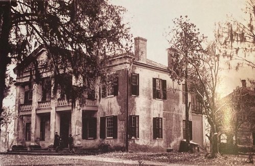 Historic two-story house with columns, surrounded by trees and a grassy area, in a black-and-white photograph.