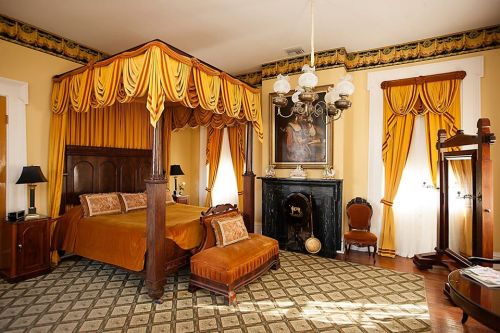 A vintage bedroom with a four-poster bed, ornate curtains, a chandelier, and a decorative fireplace.