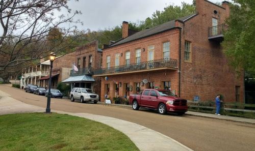 Historic brick buildings line a street, with parked cars and a person walking along the sidewalk under a cloudy sky.