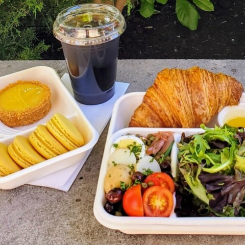 A lunch tray with a salad, croissant, and a dessert of macarons and a tart, alongside a cup of iced coffee.