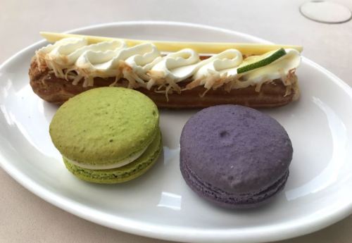 A plate with a green macaron, a purple macaron, and a coconut eclair topped with cream and a lime slice.