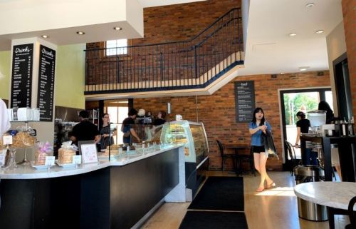 A cozy café interior with a counter displaying pastries, customers mingling, and a staircase in the background.