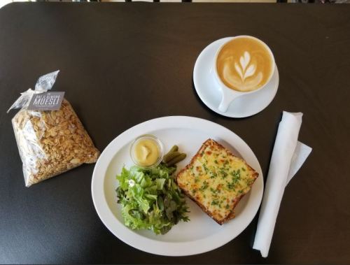 A plate with a salad, a slice of cheesy baked dish, pickles, and a cup of latte with latte art, alongside a bag of muesli.