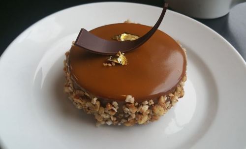 A round dessert with a glossy caramel top, surrounded by a crunchy nut crust, garnished with chocolate curls and gold leaf.