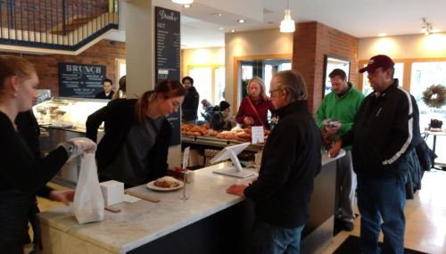 A busy café scene with customers at the counter, a barista serving food, and people waiting in line.