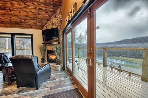 Cozy interior with a stone fireplace, large windows, and a view of a deck overlooking a scenic landscape.
