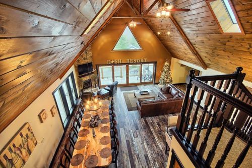 Cozy lodge interior with wooden beams, a dining table, leather sofa, and a Christmas tree by large windows.