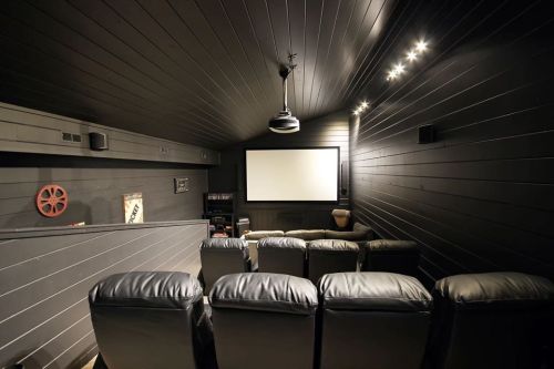 A cozy home theater with black walls, leather seating, and a large screen, illuminated by soft ceiling lights.