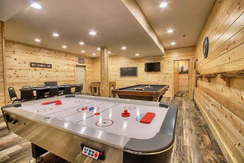 A cozy game room featuring an air hockey table, pool table, and wooden walls, with seating and a TV in the background.