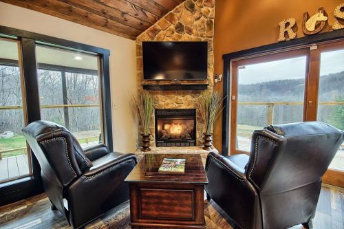 Cozy living room with two leather chairs, a stone fireplace, and a TV, overlooking a scenic outdoor view.