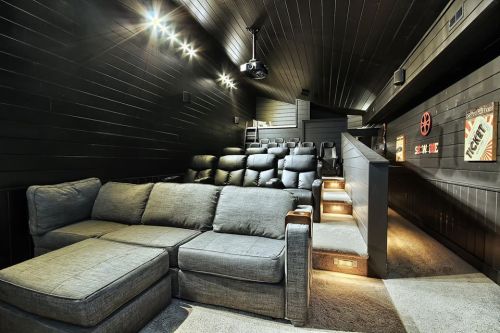 A cozy home theater with a sectional sofa, tiered seating, and dim lighting, featuring a projector and movie posters.