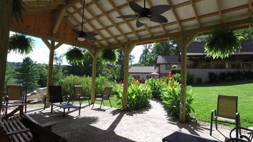 A covered patio with seating, surrounded by lush greenery and vibrant flowers, overlooking a serene landscape.