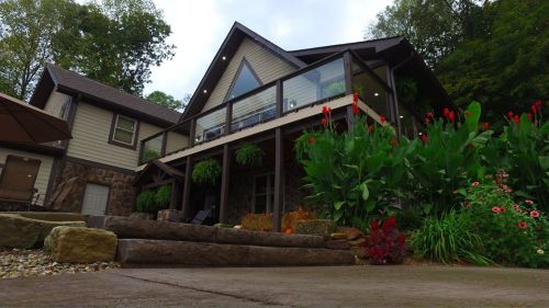 A modern house with a large deck, surrounded by lush greenery and colorful flowers, set against a cloudy sky.