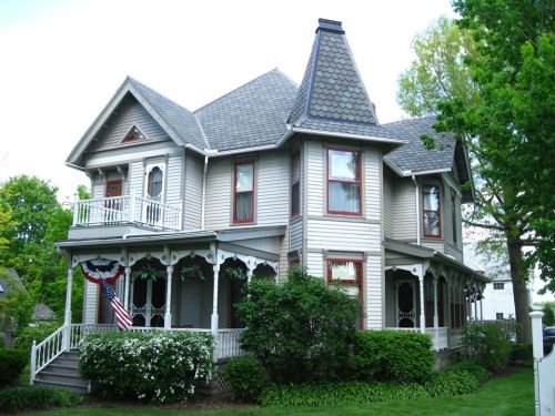 Victorian-style house with a steep roof, decorative trim, and a porch adorned with greenery and an American flag.