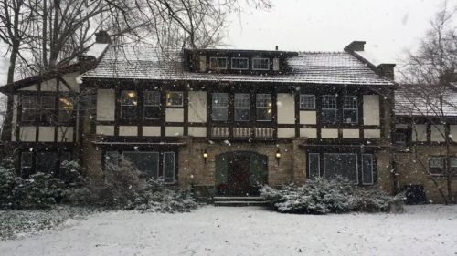 A large, historic house with a mix of stone and wood, surrounded by snow-covered trees and falling snowflakes.