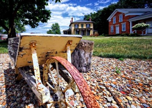 A rusty wheelbarrow sits on a gravel path, with a historic house and a red brick building in the background under a blue sky.