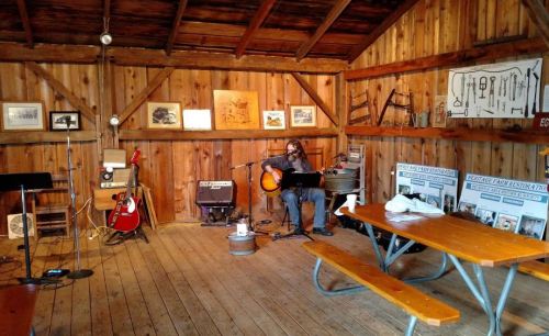 A musician plays guitar in a rustic wooden room filled with vintage photos and instruments.