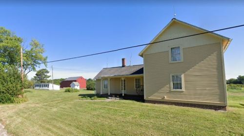 A yellow two-story house with a porch, surrounded by green grass, with a red barn and another building in the background.