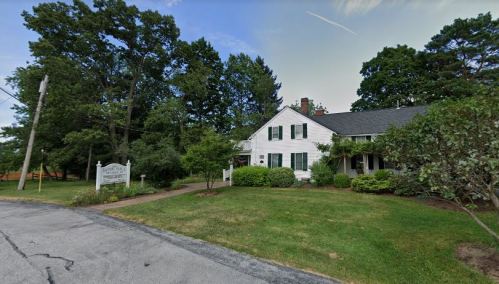 A white house with green shutters surrounded by trees and shrubs, located on a quiet street.