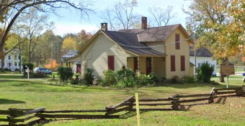 A yellow house with red shutters surrounded by greenery and a wooden fence, set in a rural landscape.