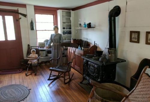 A vintage room with wooden floors, a rocking chair, a stove, and mannequins dressed in historical clothing.