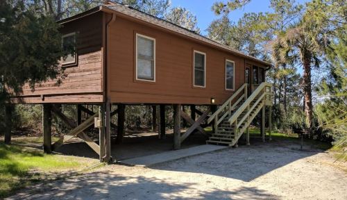 A wooden house on stilts surrounded by trees, featuring a staircase and a gravel path leading to the entrance.
