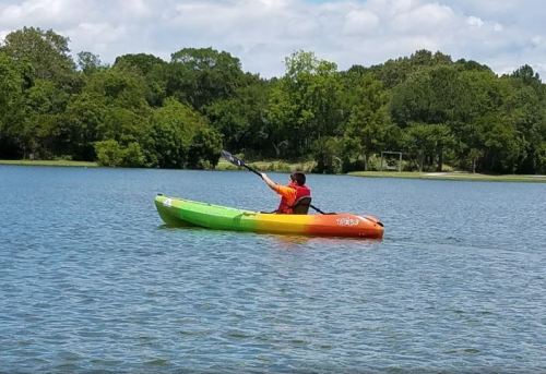 A person in a colorful kayak paddles on a calm lake surrounded by greenery under a partly cloudy sky.