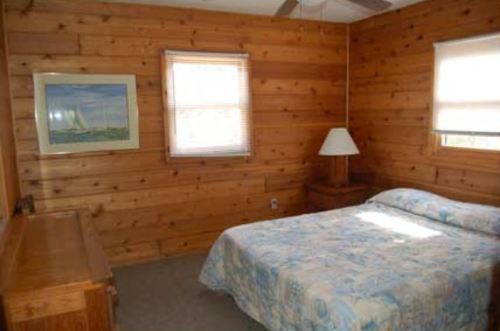 Cozy bedroom with wooden walls, a double bed, lamp, dresser, and a window with blinds.