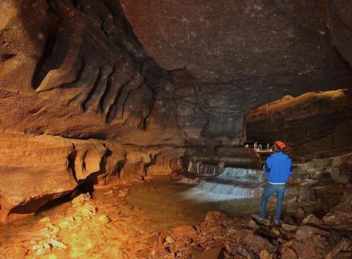 A person in a blue jacket stands in a large cave with rock formations and a small stream of water.