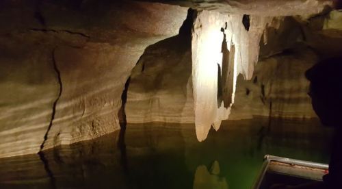 A dimly lit cave with water reflecting rock formations and a person in a boat on the right side.