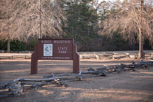 Sign for Kings Mountain State Park, surrounded by trees and a wooden fence, in a grassy area.