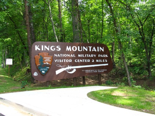 Sign for Kings Mountain National Military Park, indicating the visitor center is 2 miles ahead, surrounded by trees.