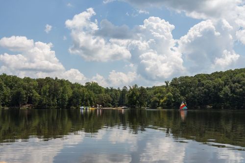 A serene lake surrounded by lush greenery, with colorful sailboats and fluffy clouds reflected on the water's surface.