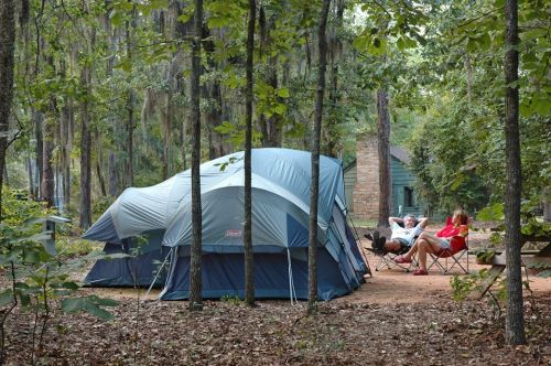 A blue tent in a wooded area with two people relaxing in chairs nearby, surrounded by trees and nature.