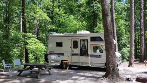 A camper parked in a wooded area, surrounded by trees, with a picnic table and chairs nearby.